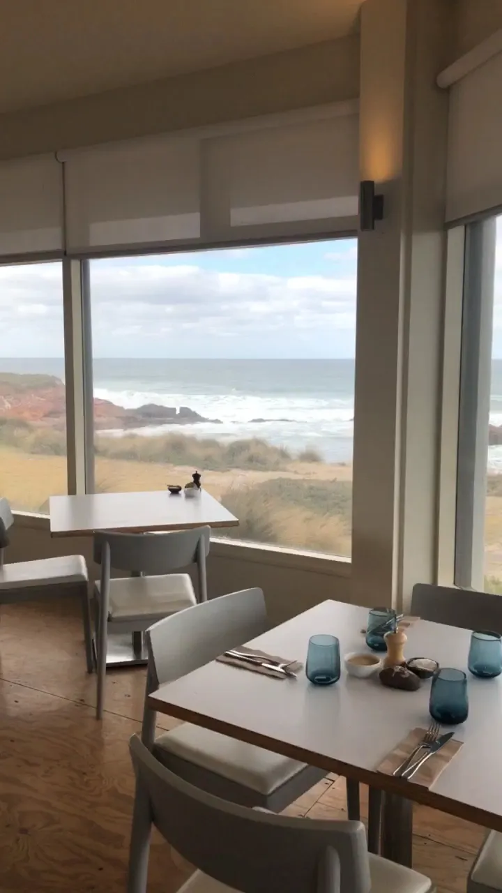Empty Cape Kitchen dining tables beside large windows overlooking a wild grey winter ocean